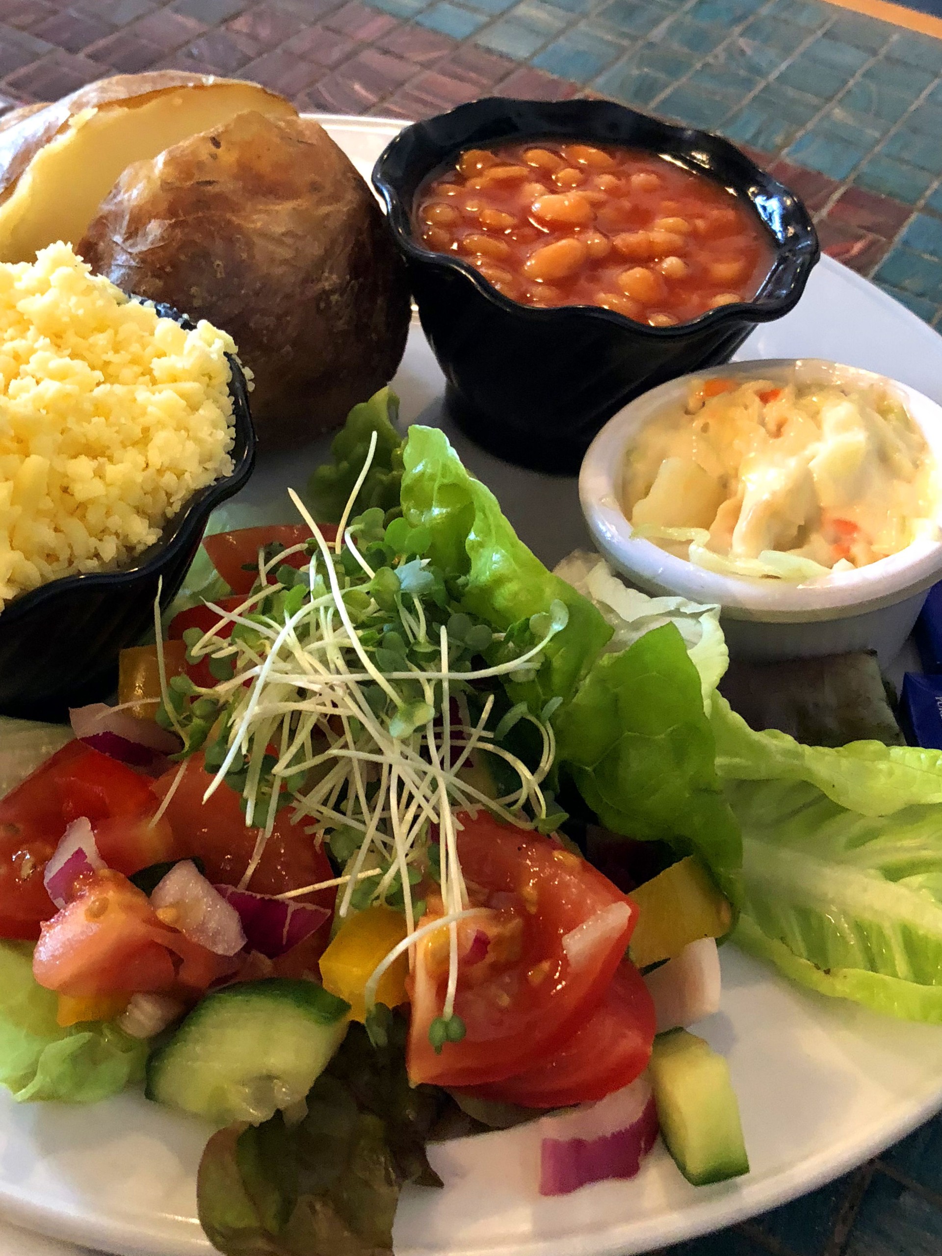 Image of baked potato with grated cheese, baked beans, coleslaw and salad, eating out jacket potatoes pub grub food served with lettuce leaves, cucumber, tomatoes, mixed bell peppers and chopped cress garnish, five a day healthy eating high fibre diet